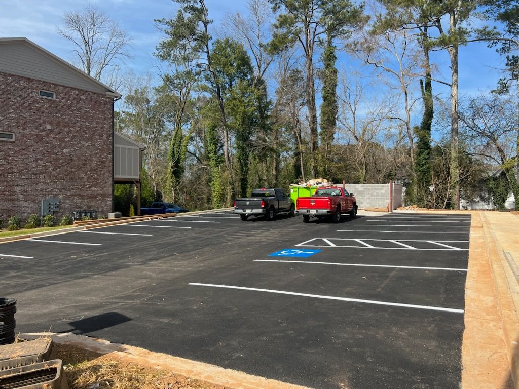 Newly paved parking lot with marked spaces. Two trucks parked. Building on left, trees in the background.
