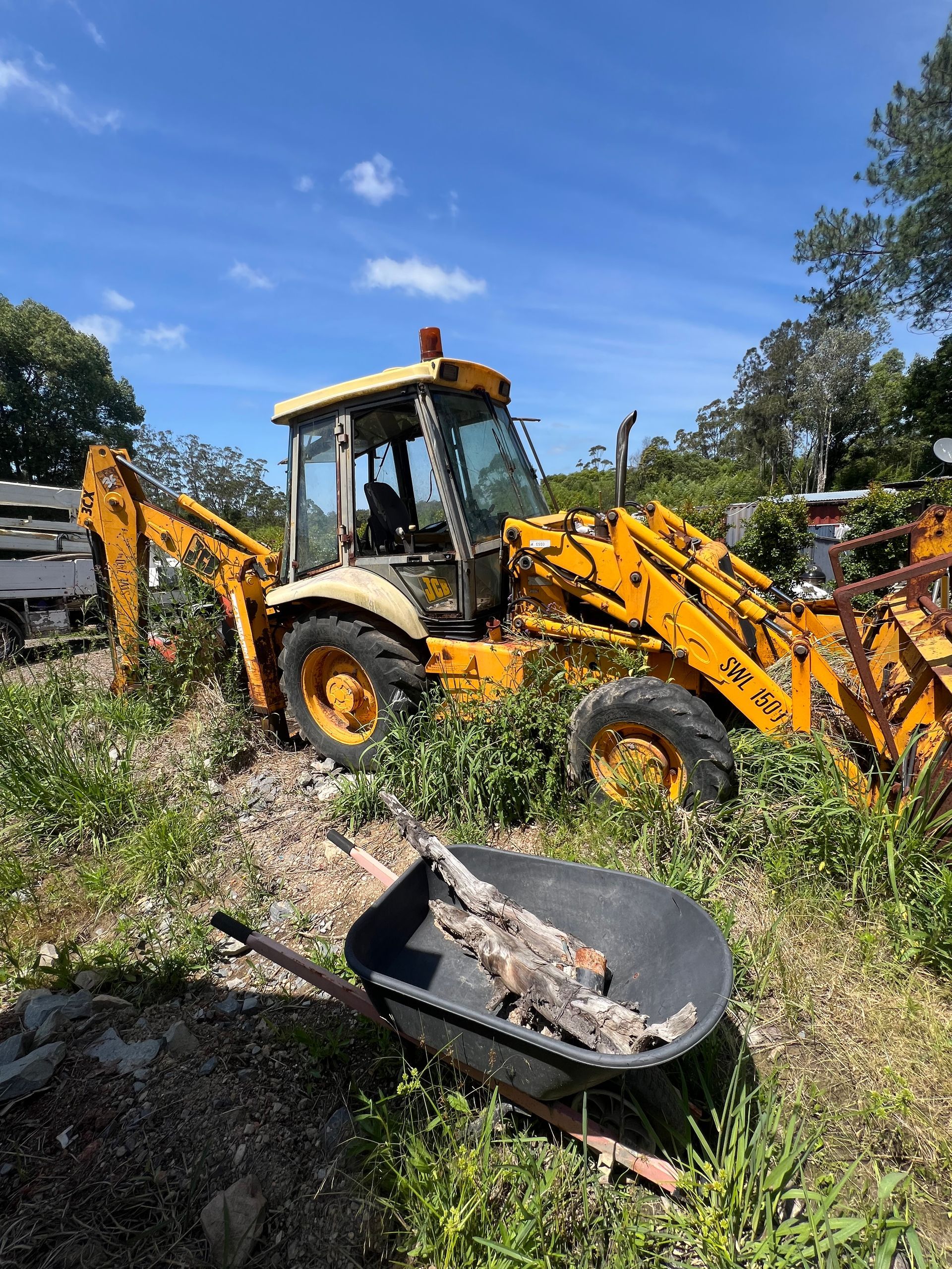 Yellow Backhoe Overgrown With Weeds, Wheelbarrow in Foreground — North Coast Heavy Diesel In Kempsey, NSW