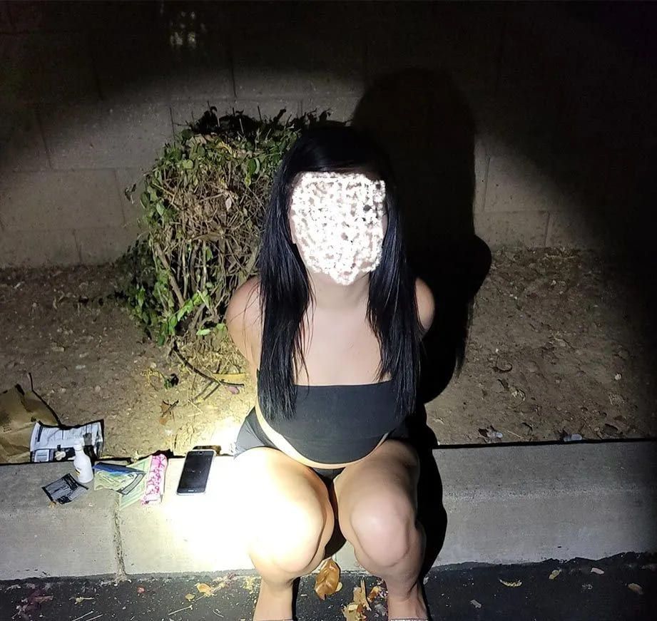 A woman is kneeling down on the sidewalk with a flashlight on her face.