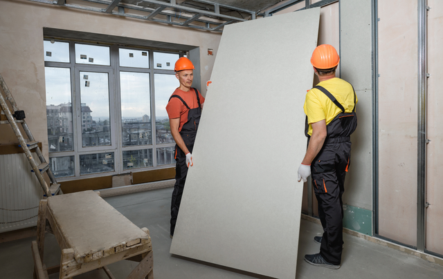 Two construction workers are carrying a large piece of drywall in a room.