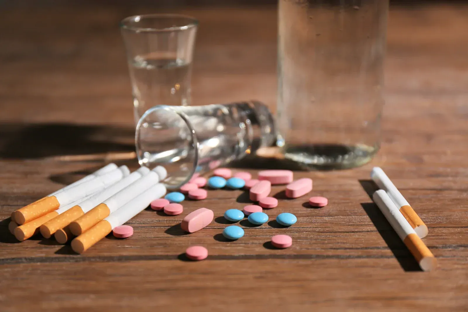 Cigarettes , pills , a glass of water and a shot glass on a wooden table.