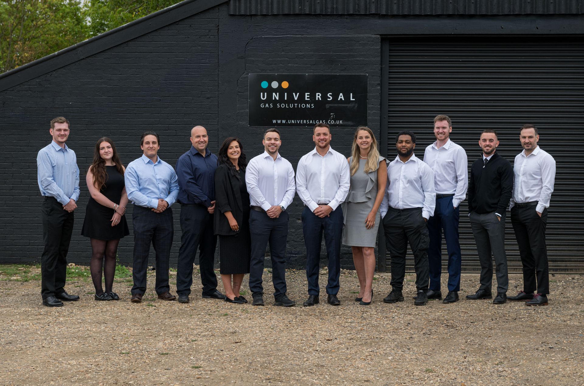 Group of 13 people in business attire stand in front of a dark building with a logo.