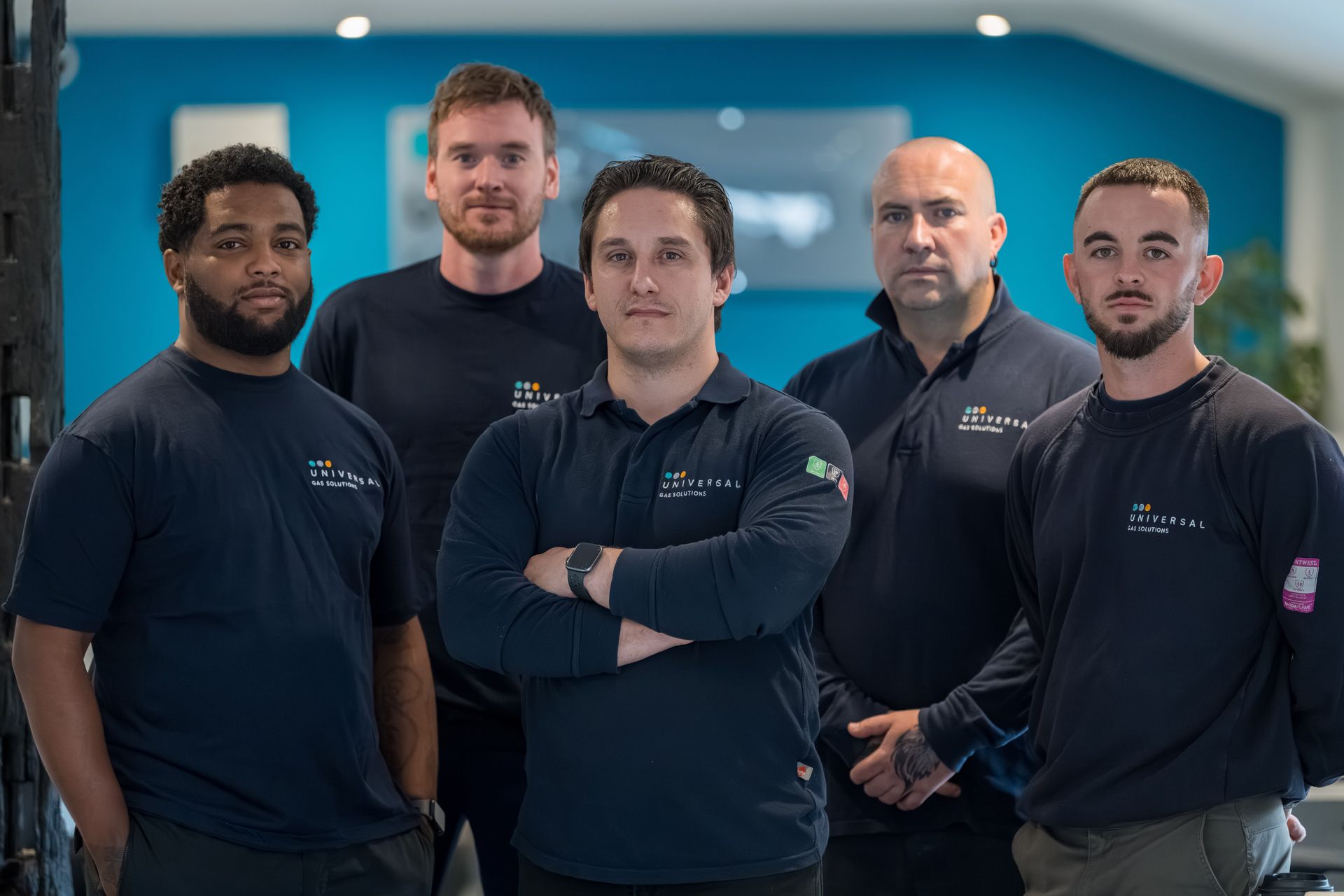 A group of five men in navy shirts posing. Indoor setting with blue wall, serious expressions.