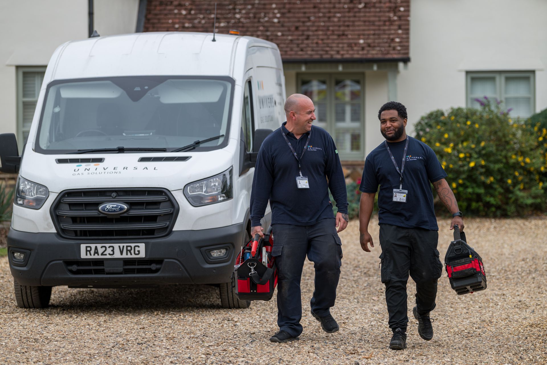 Two men in work uniforms with tool bags, walking toward a house next to a white van.