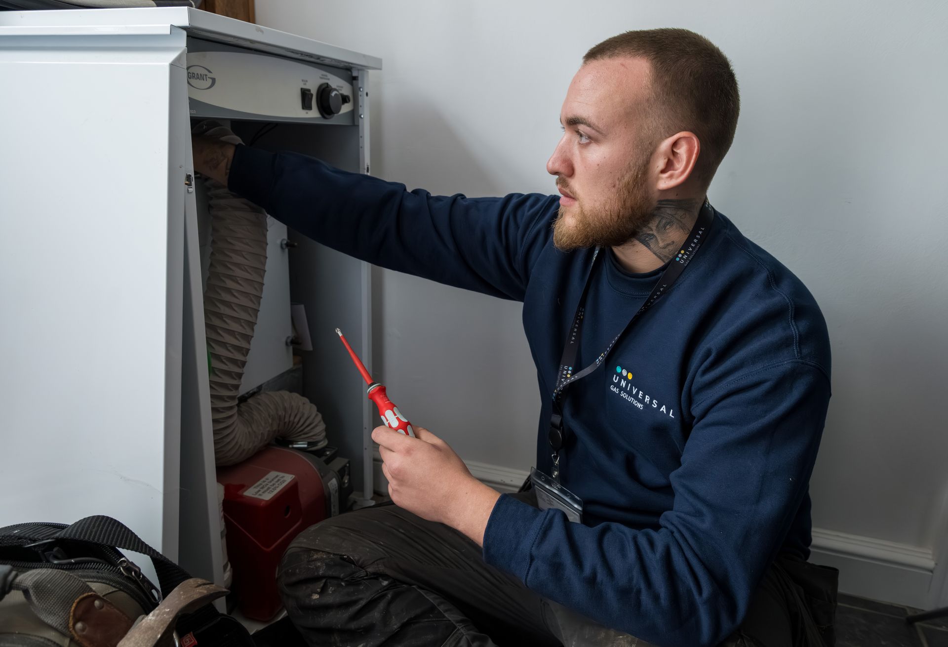 A man in blue sweater examines a boiler with a screwdriver. Indoors, light-skinned, with tattoos.