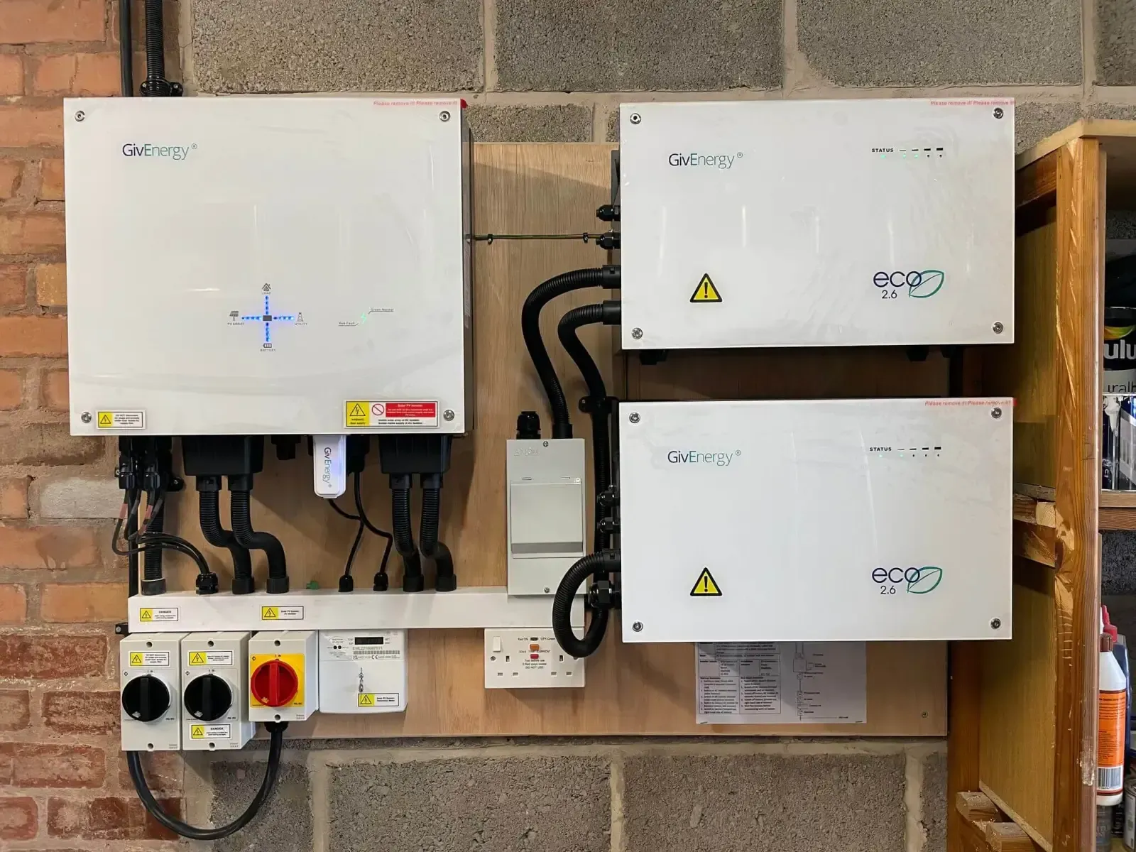 Solar panel inverters and electrical components mounted on wooden board against brick wall.
