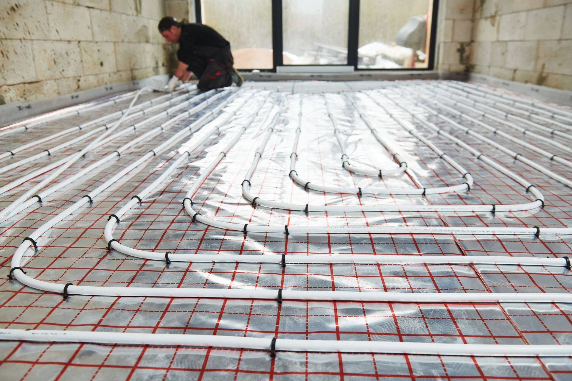 Person installing radiant floor heating system: white tubing on red grid in room.