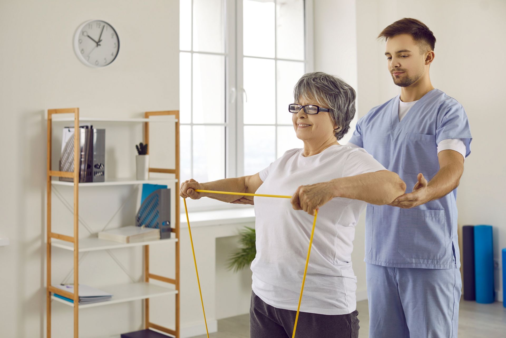 A senior lady is doing exercises with rubber, with a physiotherapist standing behind and helping her.