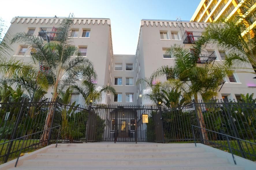 Gray apartment building behind a black iron fence; palm trees and stairs in front.