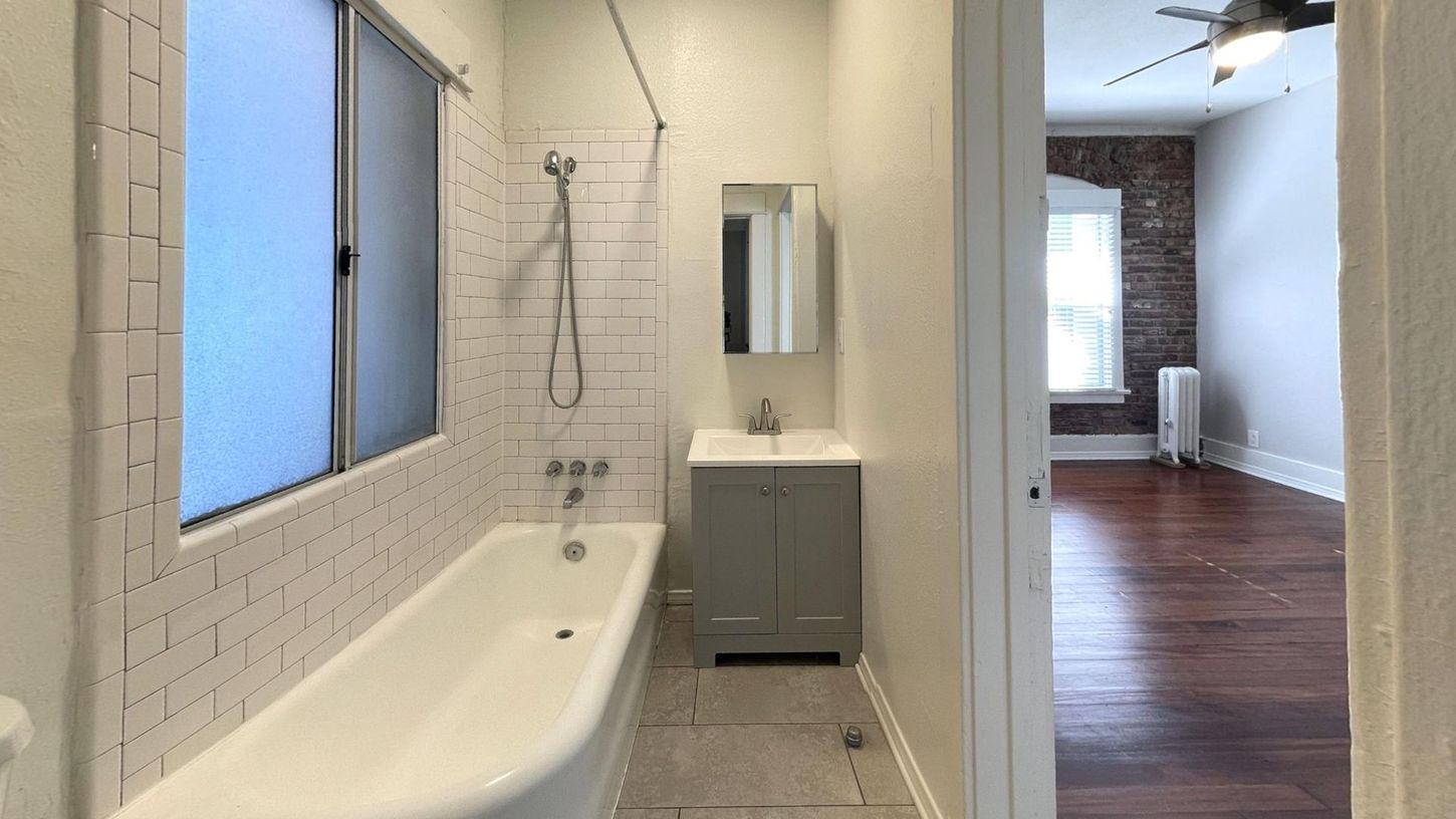 Bathroom with white bathtub, subway tile, and grey vanity, doorway to a room with hardwood floors and exposed brick.