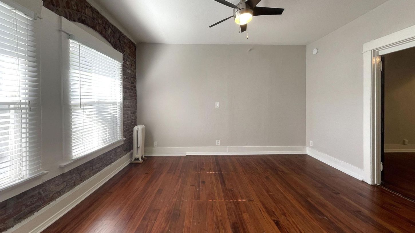 Empty room with hardwood floors, exposed brick wall, window with blinds, and ceiling fan.