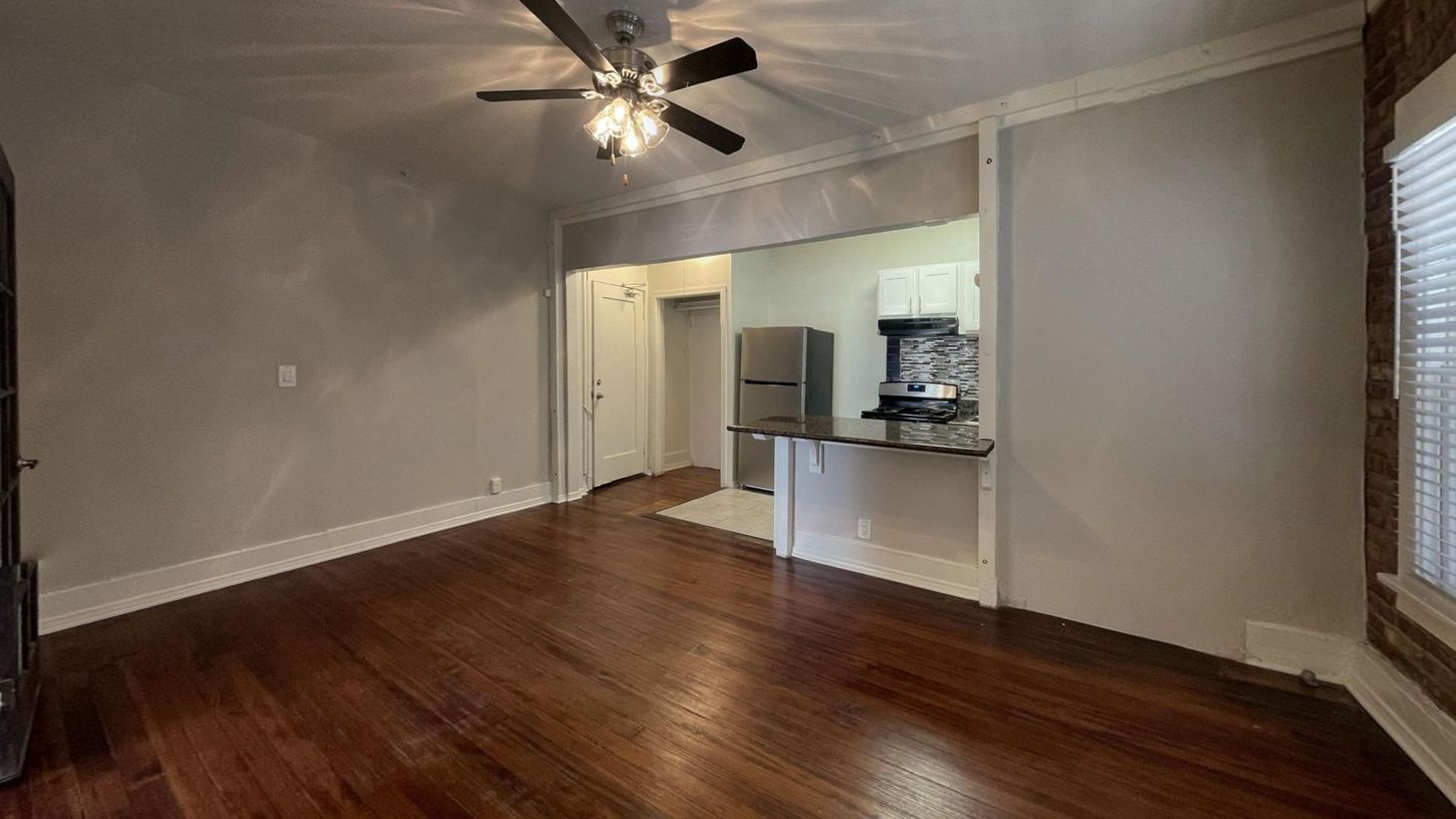 Empty apartment living space with dark wood floors, open to kitchen, brick wall, and a ceiling fan.
