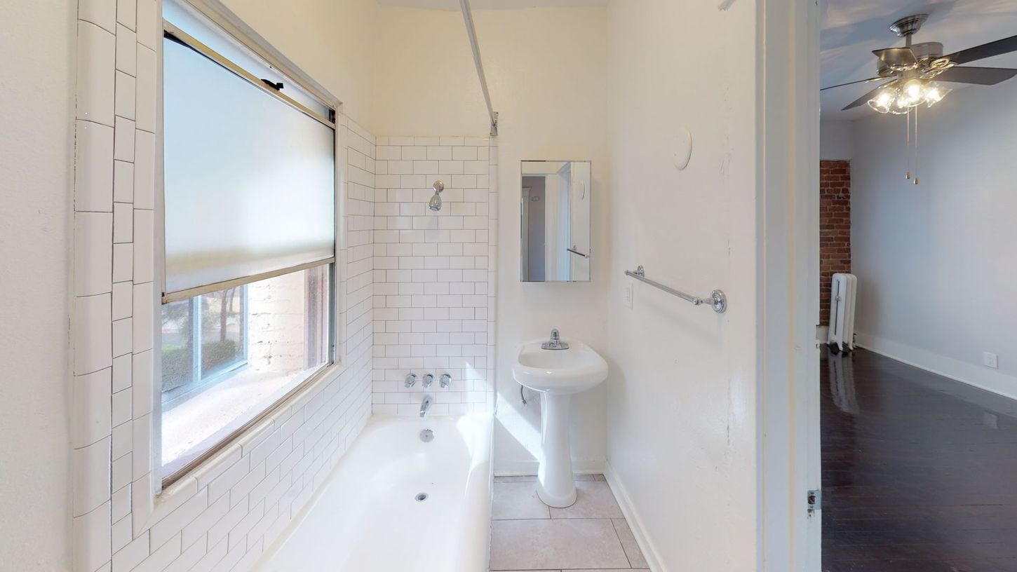 Bathroom with bathtub, sink, window, and mirror. White walls and brick accents. Dark wood floor visible through doorway.