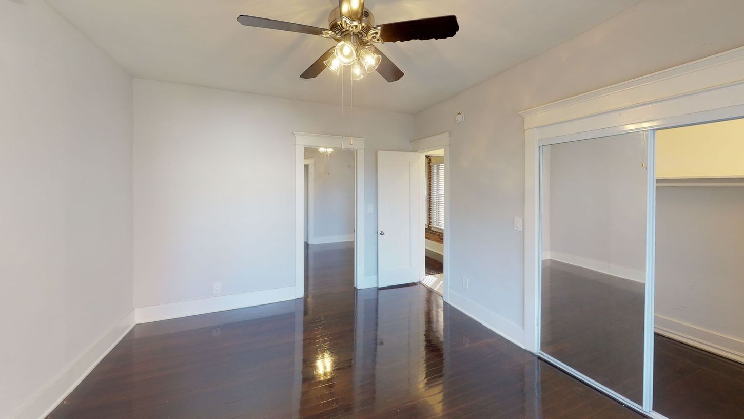 Empty bedroom with hardwood floors, white walls, and a ceiling fan. Open closet and doorways.
