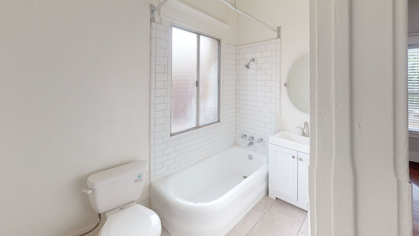 Bathroom with white walls, tub, toilet, and vanity with a round mirror. Window has frosted glass.