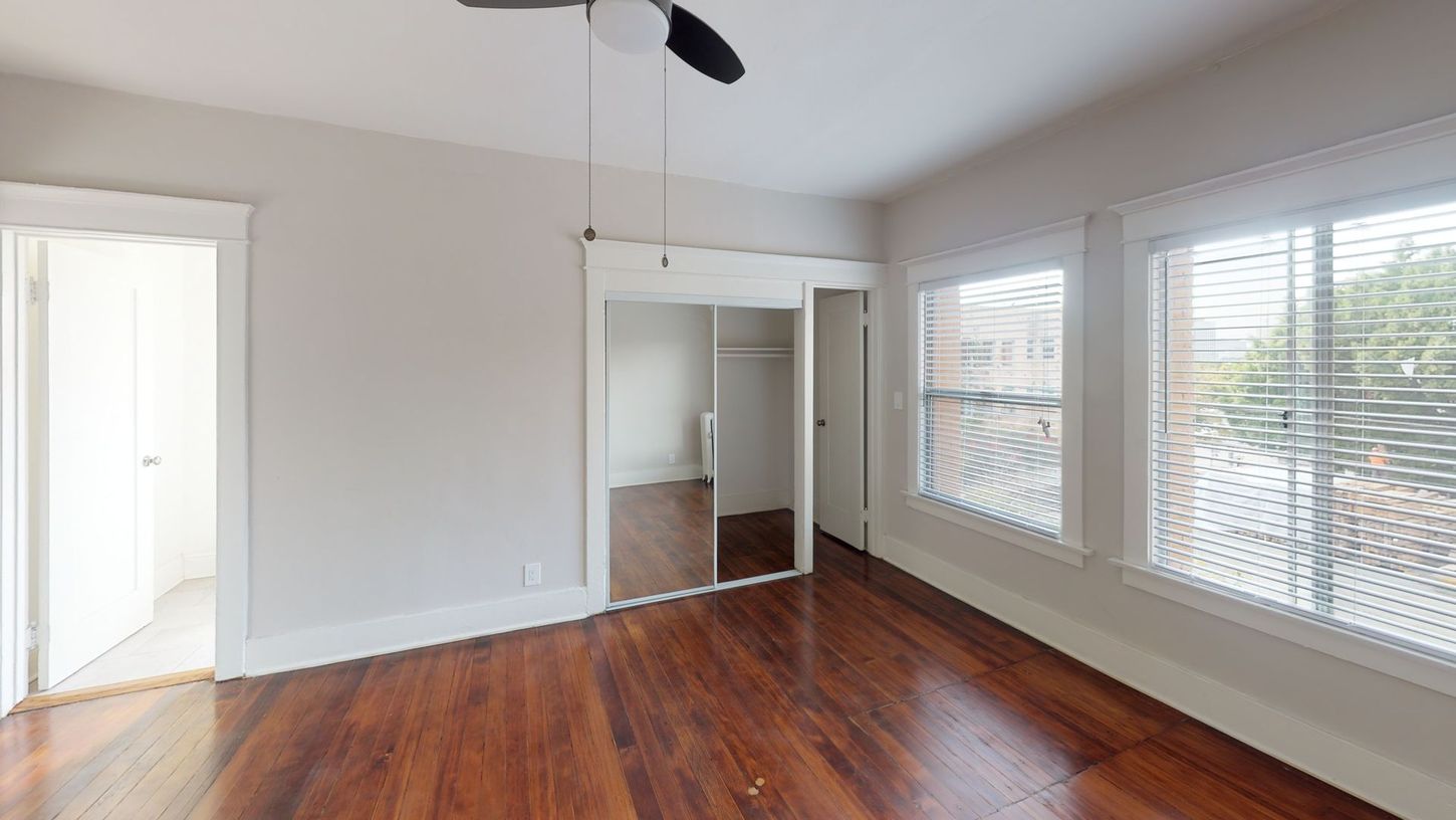 Empty room with hardwood floors, closet, windows with blinds, and ceiling fan.