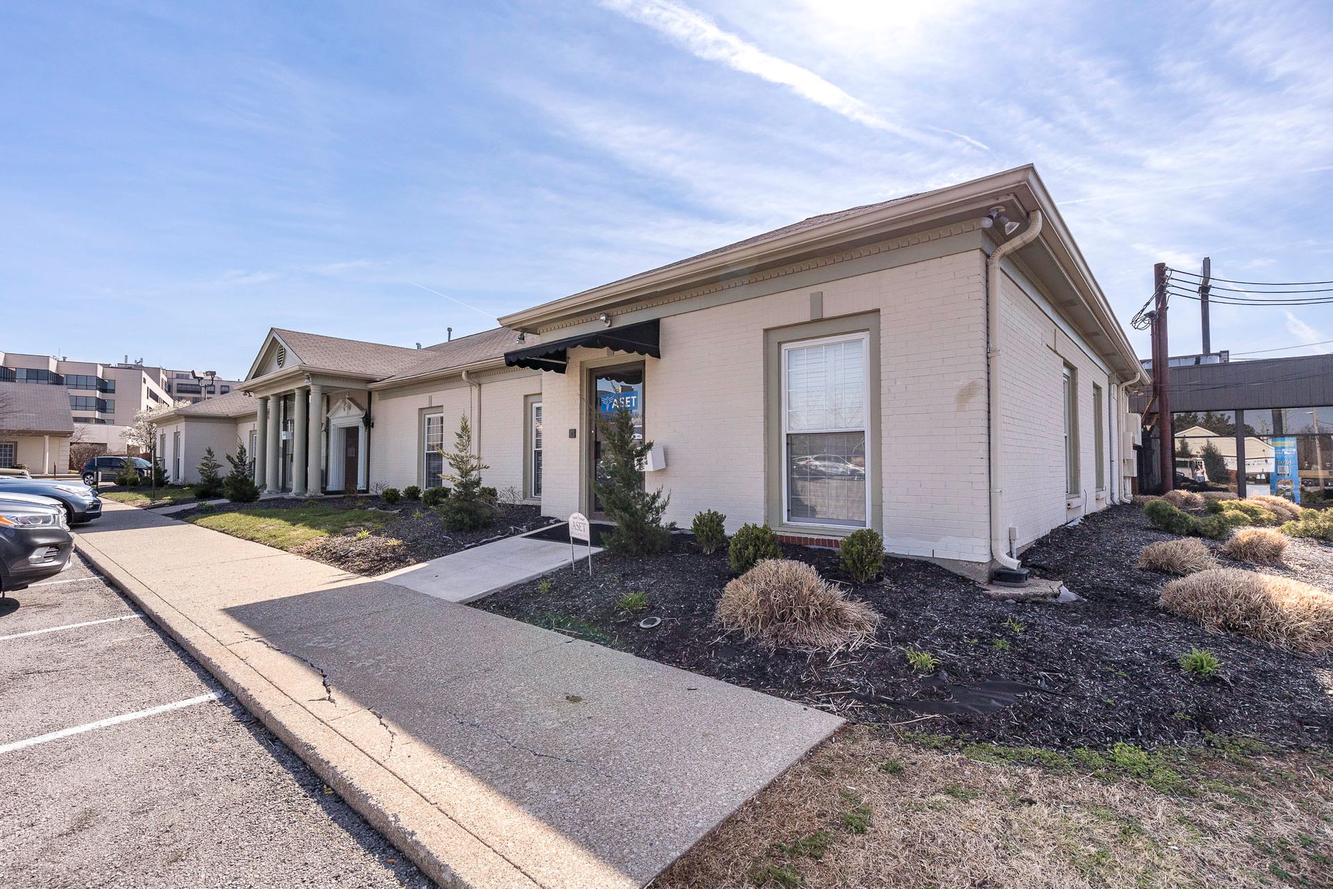 A small white building with a lot of windows is sitting next to a parking lot.