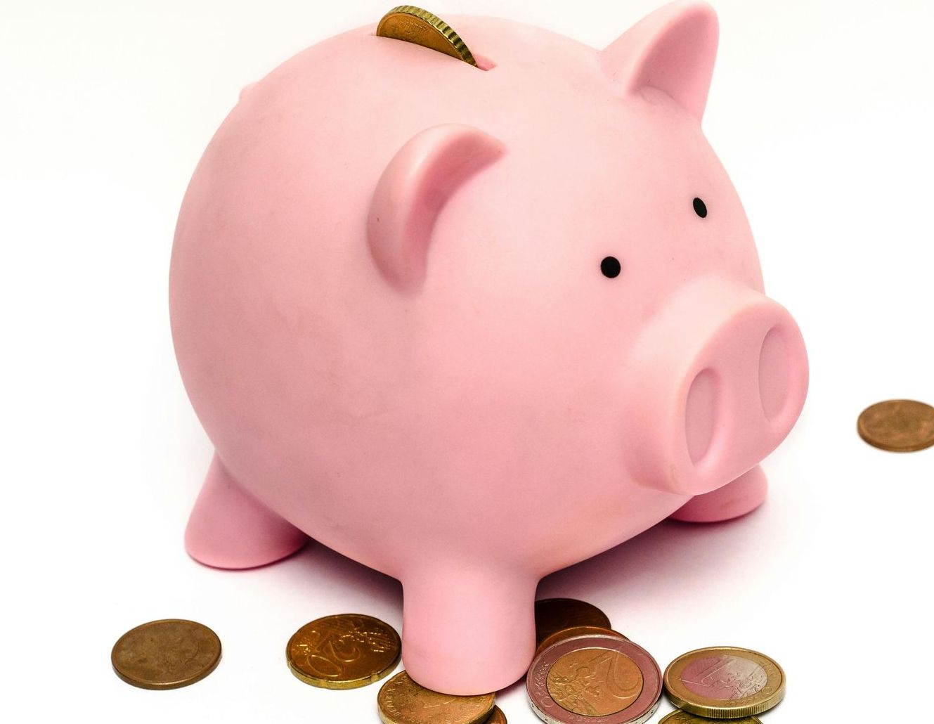 Pink piggy bank with a coin in the slot, surrounded by loose coins on a white surface.