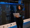 A woman with dark curly hair stands in a server room, looking at a tablet. She wears a black sweater and pants.