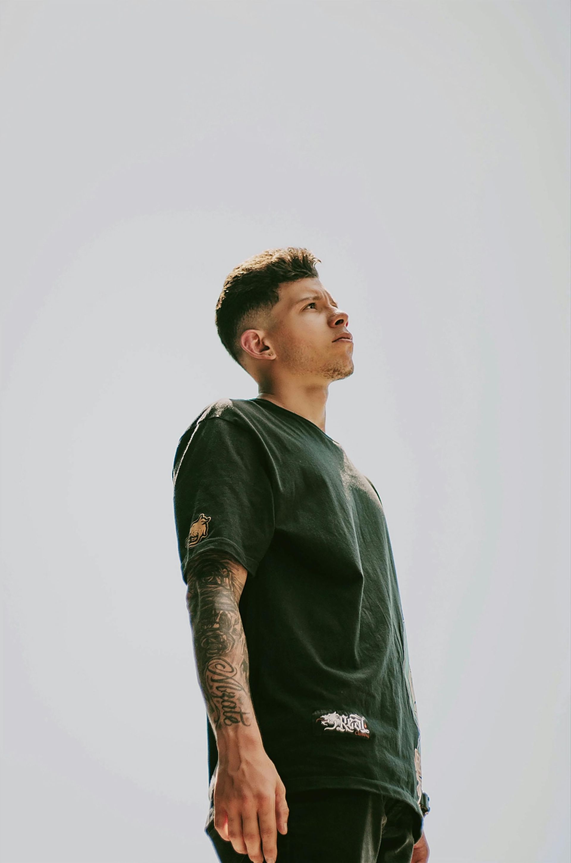 Man with a tattoo sleeve looks upward against a bright sky, wearing a dark green t-shirt.