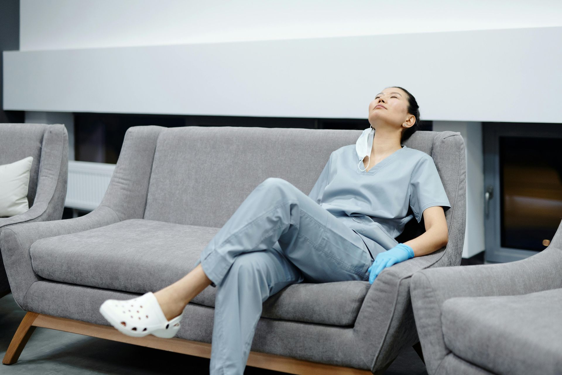 A healthcare worker in scrubs and Crocs rests on a gray sofa, eyes closed, face mask dangling.