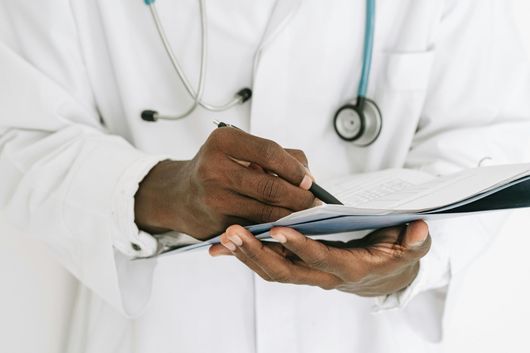 Doctor in white coat and with stethoscope, holding and writing on a clipboard.