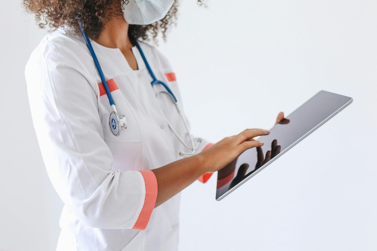 A medical professional in a white coat and mask uses a tablet computer, stethoscope around their neck.