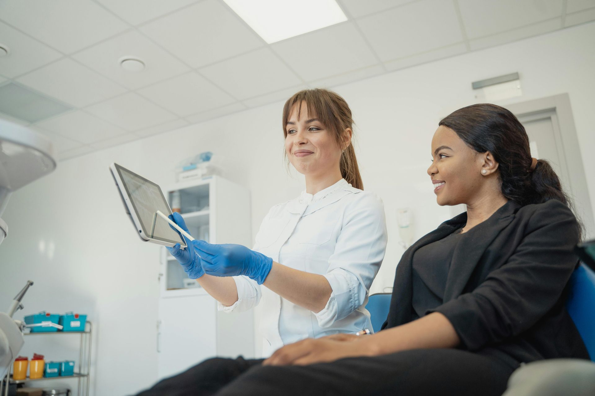 A doctor in a white coat shows an X-ray on a tablet to a smiling Black patient in a dental office.