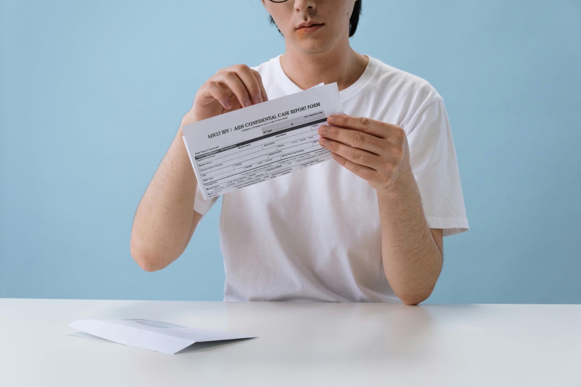 A person in a white shirt tears a document in front of a blue backdrop. A white envelope sits on a table.