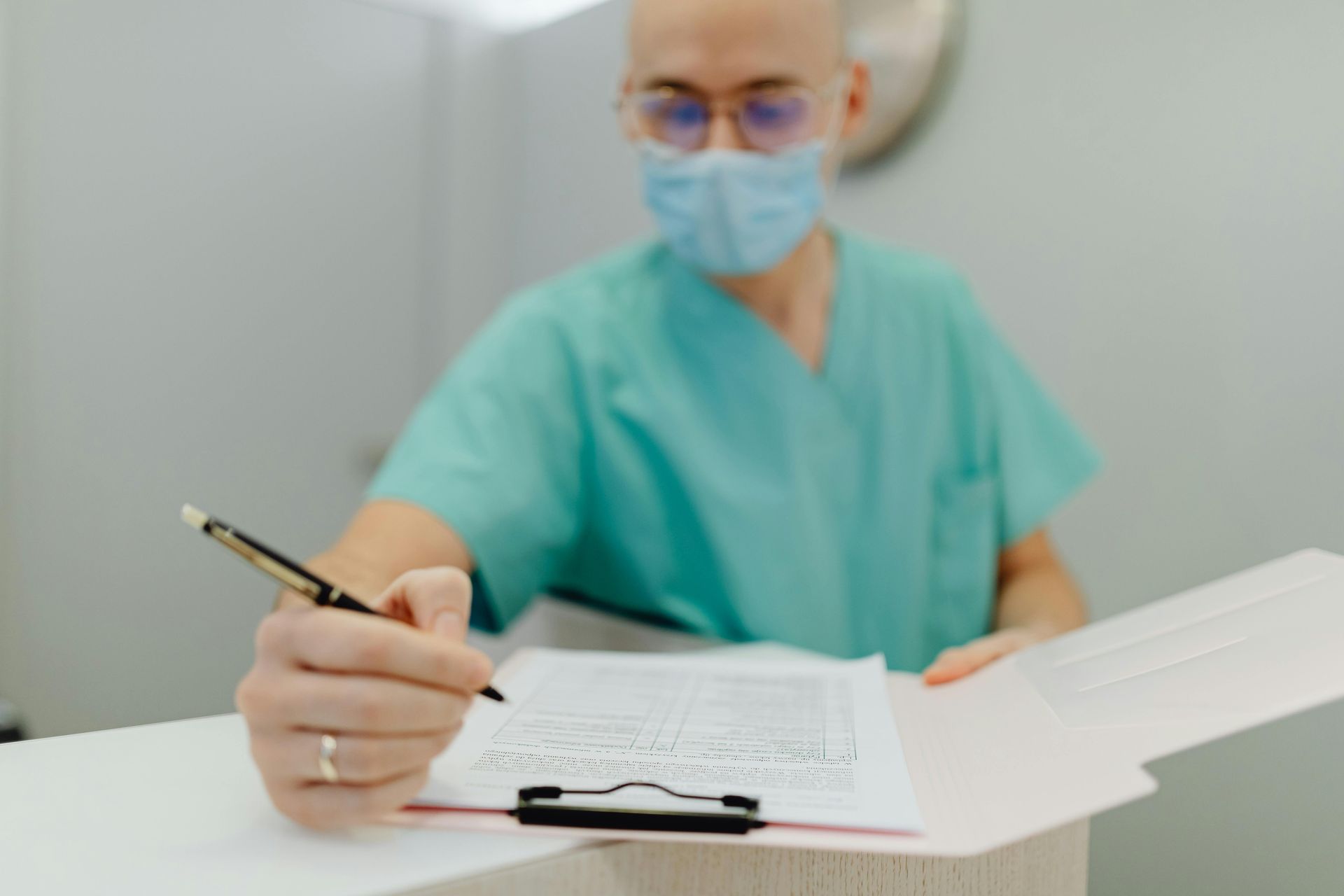Doctor in a surgical mask and scrubs writing on a clipboard at a desk.