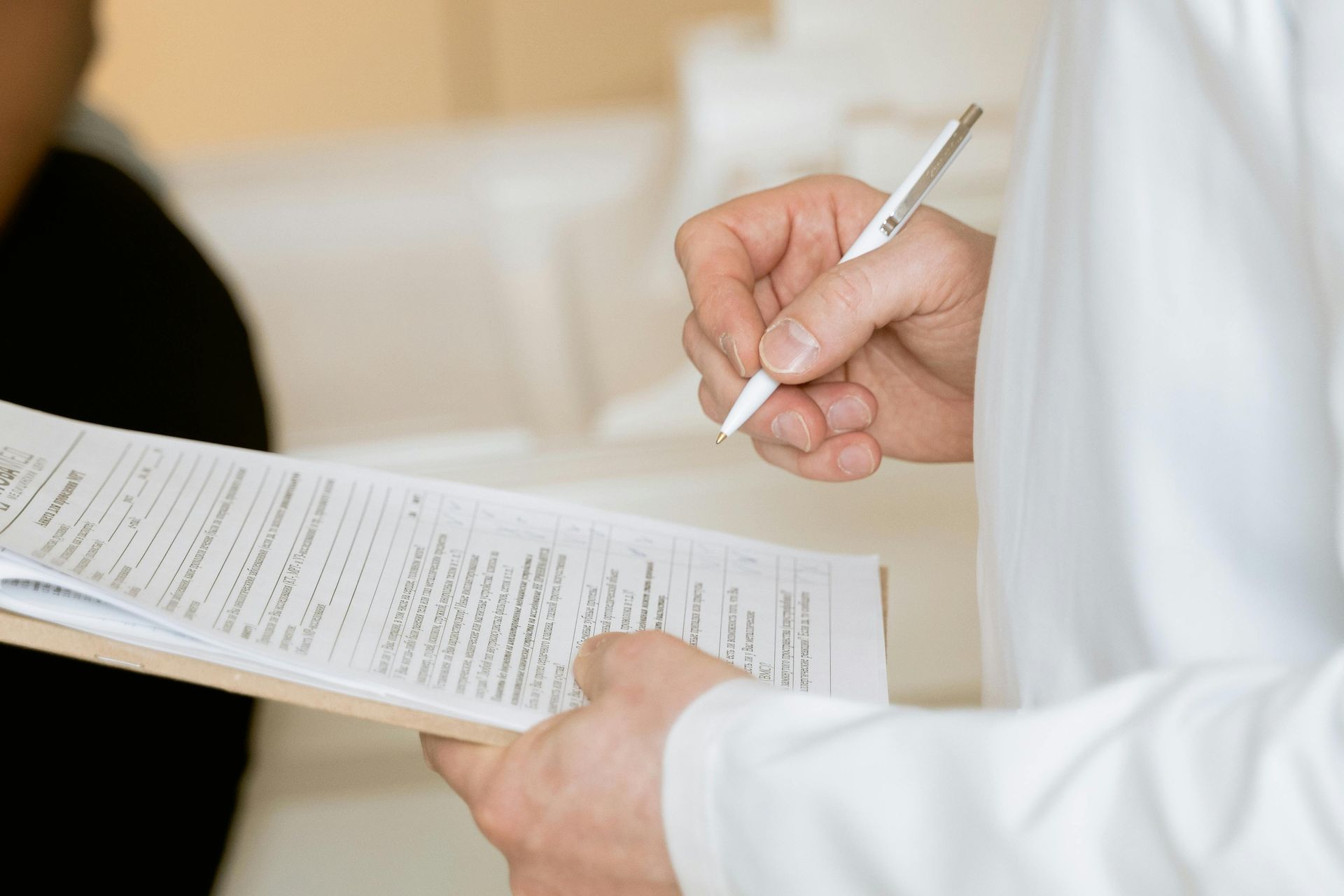 Doctor in white coat writing on medical paperwork held on a clipboard; patient out of focus on the left.