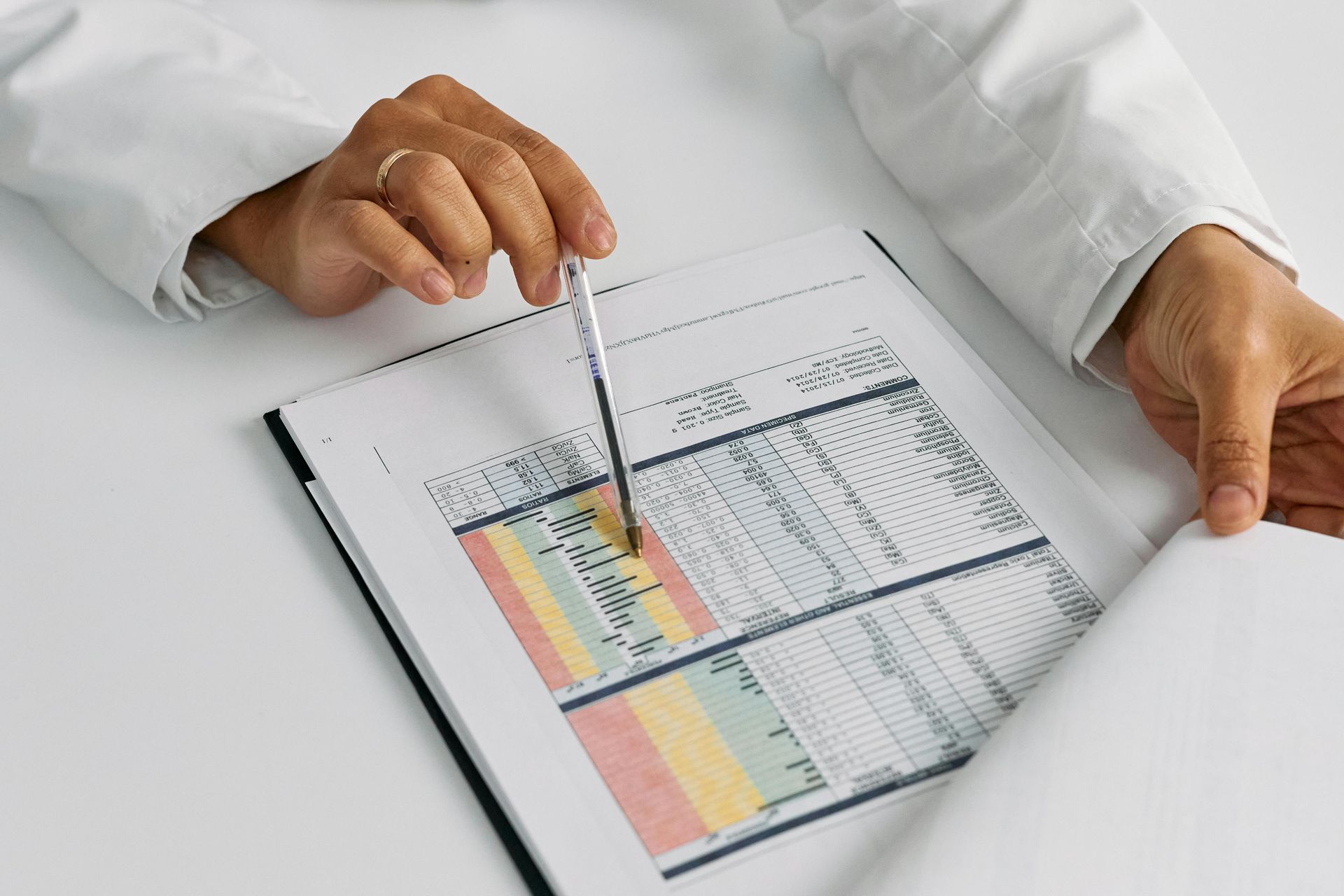 A person in a lab coat points a pen at a color-coded chart on a clipboard, likely reviewing data.