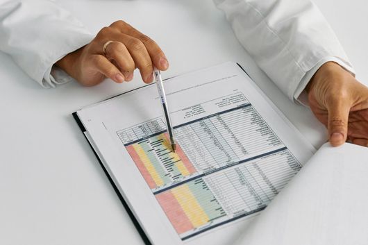 A person in a lab coat points a pen at a color-coded chart on a clipboard, likely reviewing data.