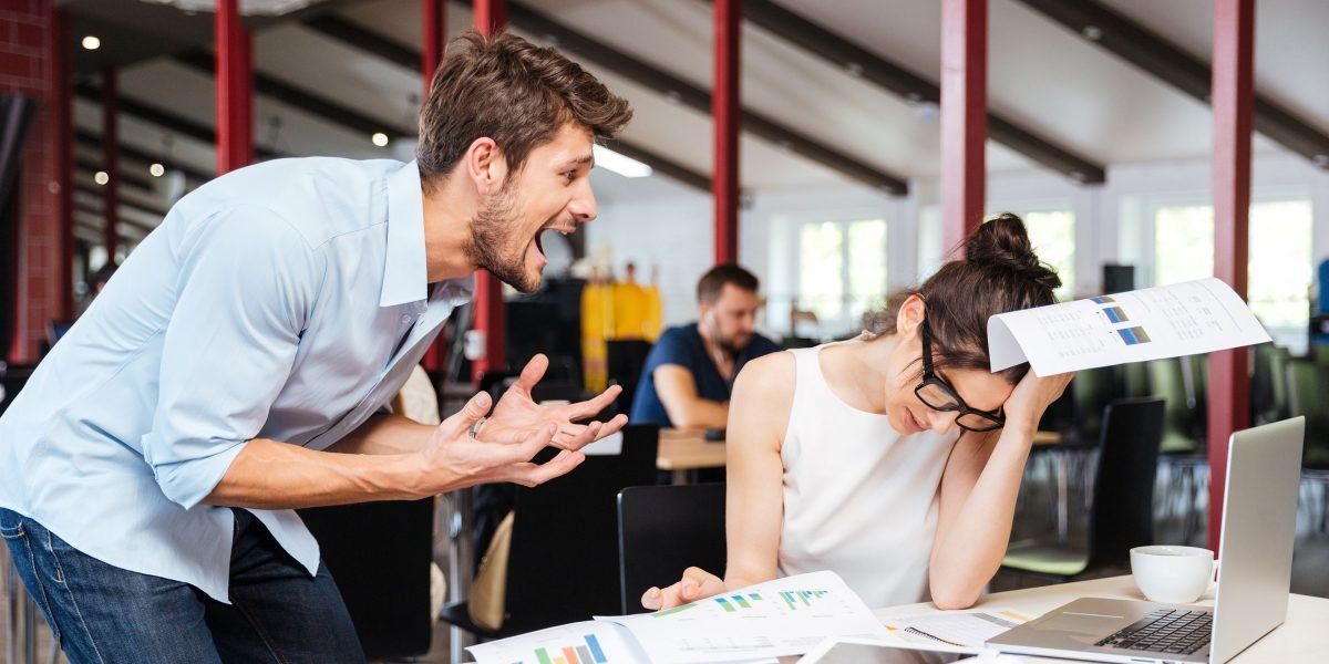 A man is yelling at a woman who is sitting at a desk with a laptop.