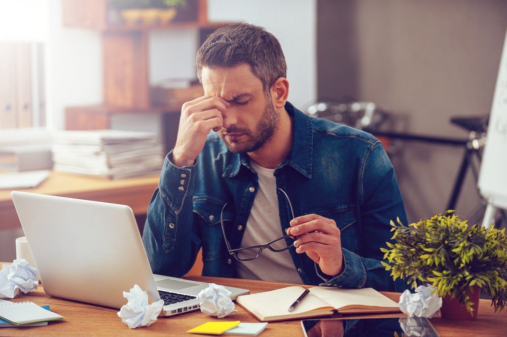 A man is sitting at a desk with a laptop and rubbing his nose.