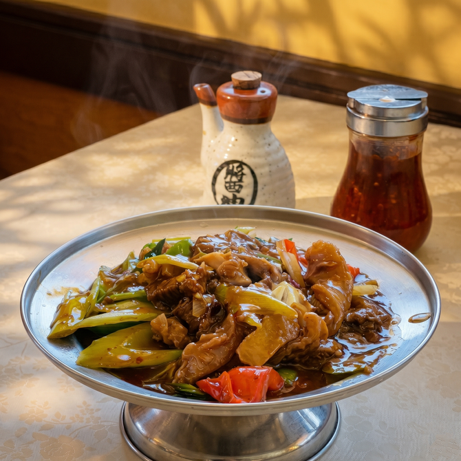 A metal bowl of stir-fried meat and vegetables sits on a table next to a soy sauce dispenser and a jar of chili oil.
