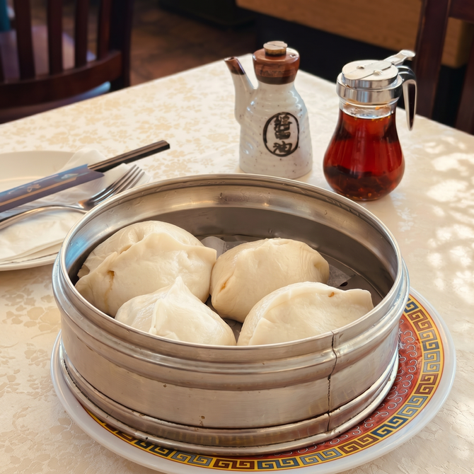 A metal steamer basket containing four steamed bao buns sits on a white table with soy sauce and chili oil dispensers.