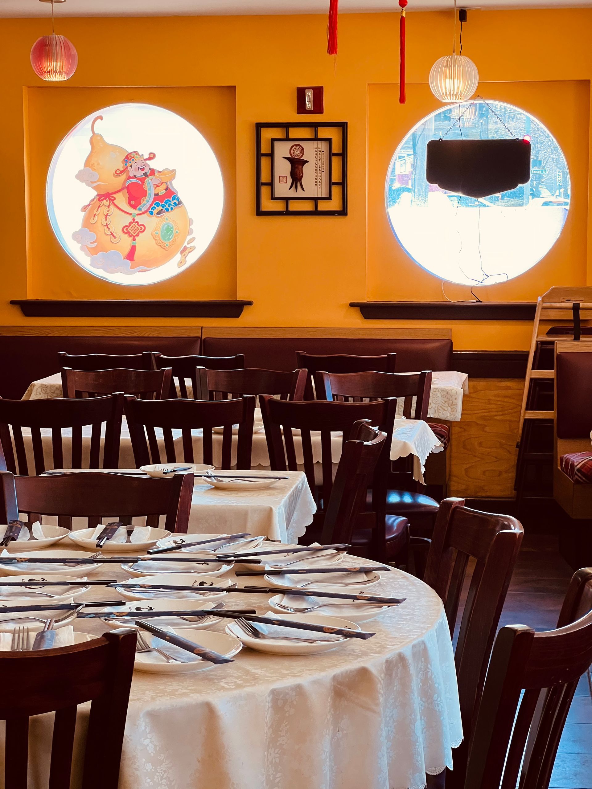 A dining room with yellow walls, round windows, and tables set with white tablecloths and dark wooden chairs.