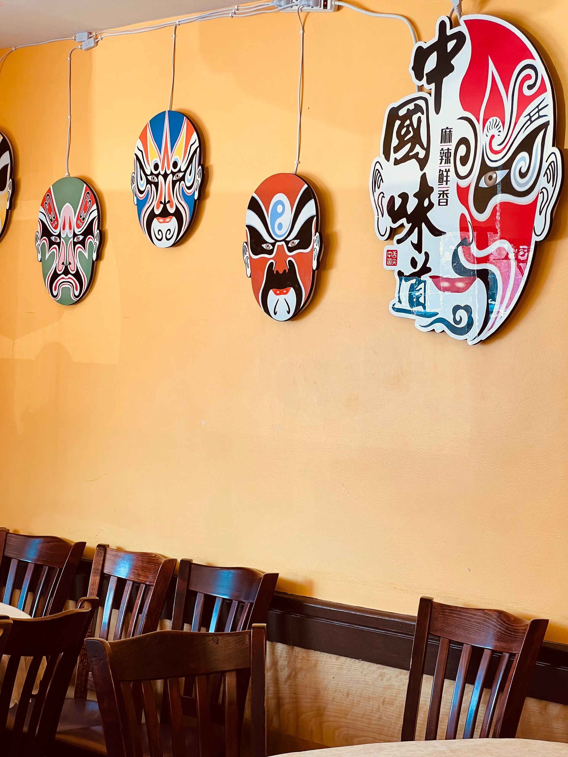A wall decorated with several hanging Chinese opera masks above wooden chairs in a restaurant.