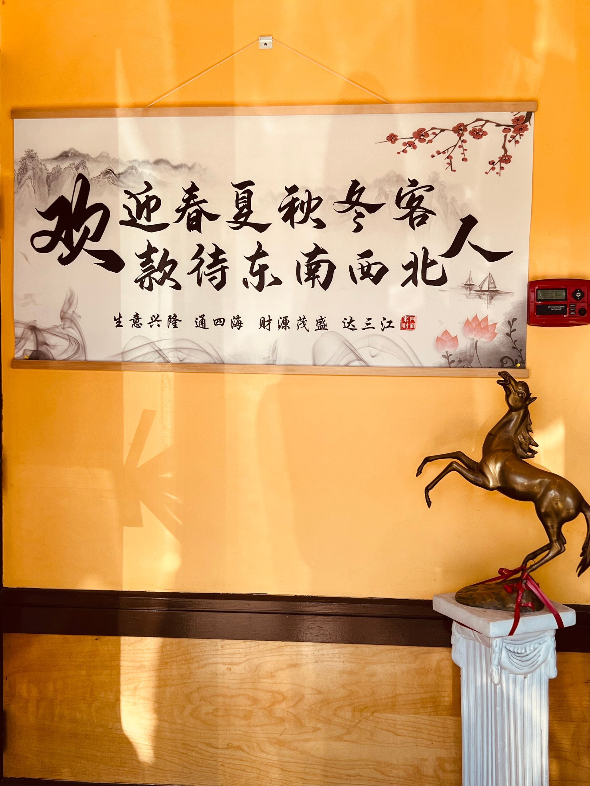 A wall hanging with Chinese calligraphy on a yellow wall next to a bronze horse statue on a pedestal.