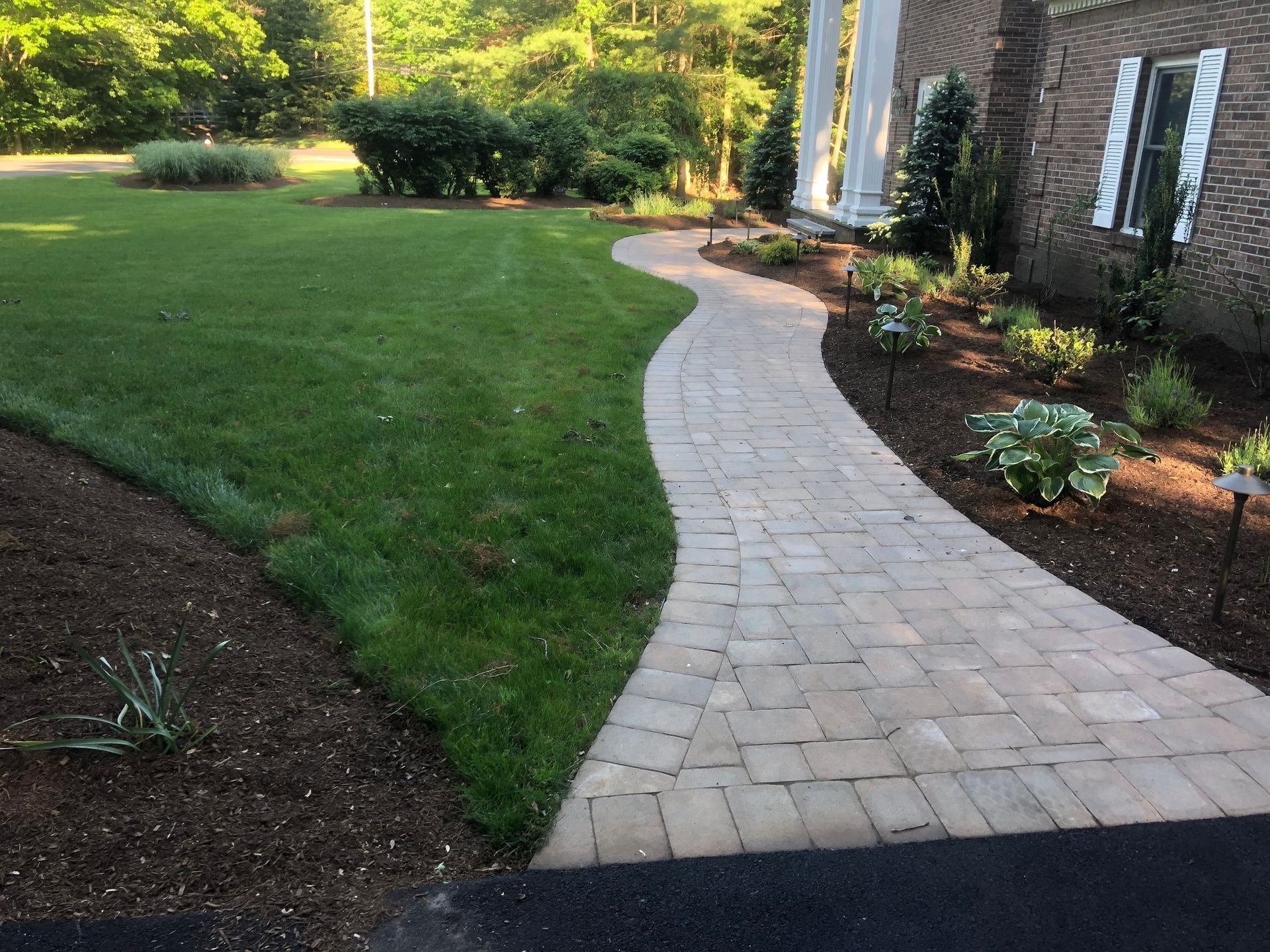 A curved stone paver walkway leads through a landscaped yard to a brick home entrance surrounded by green trees and mulch.