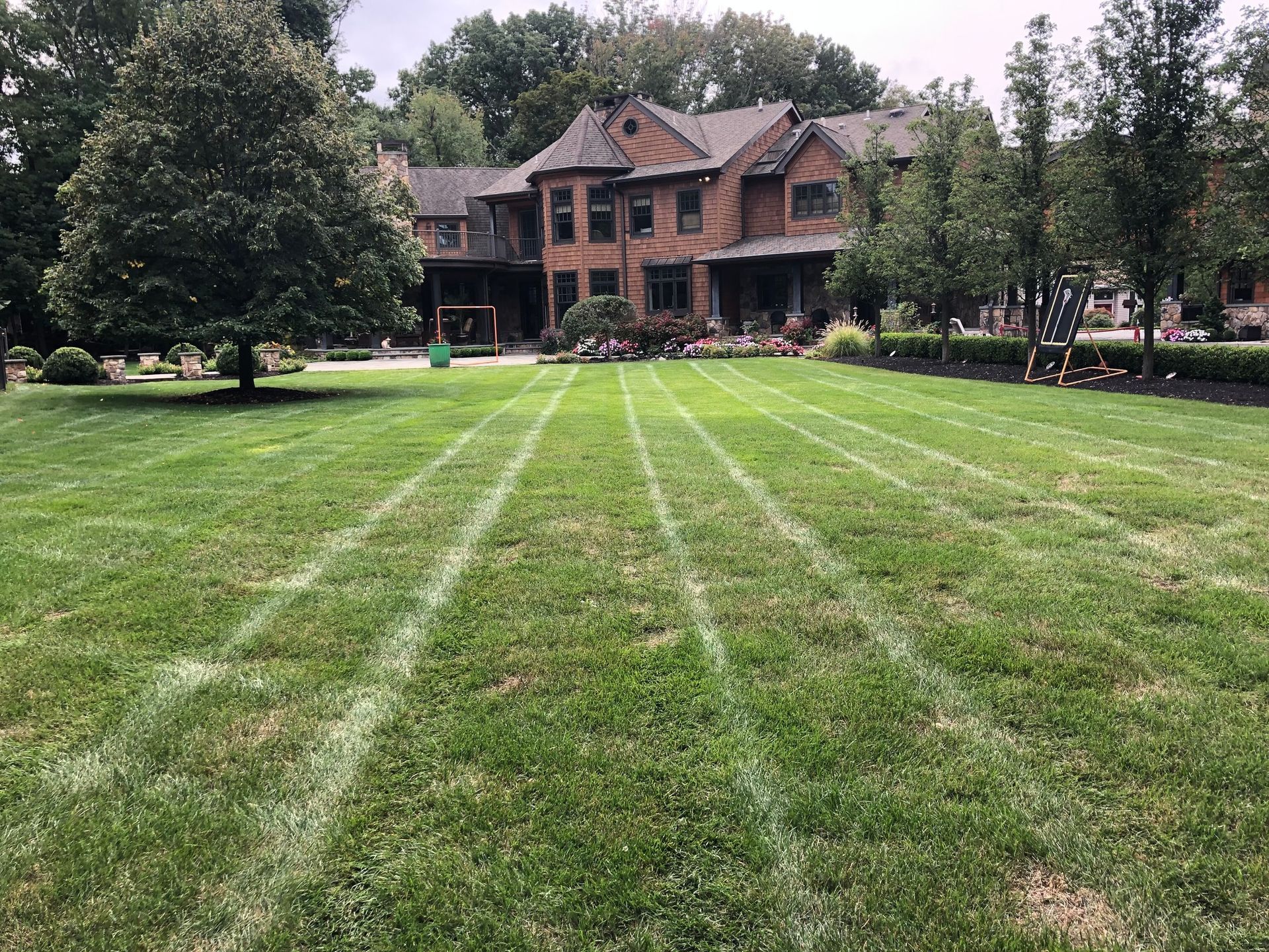 A large lawn with vertical lawnmower striping leads to a multi-story brick house surrounded by mature trees.