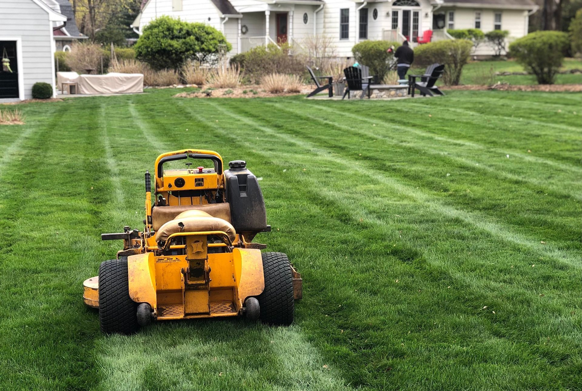 A yellow riding lawn mower sits in the middle of a large, neatly striped green lawn in front of a suburban home.