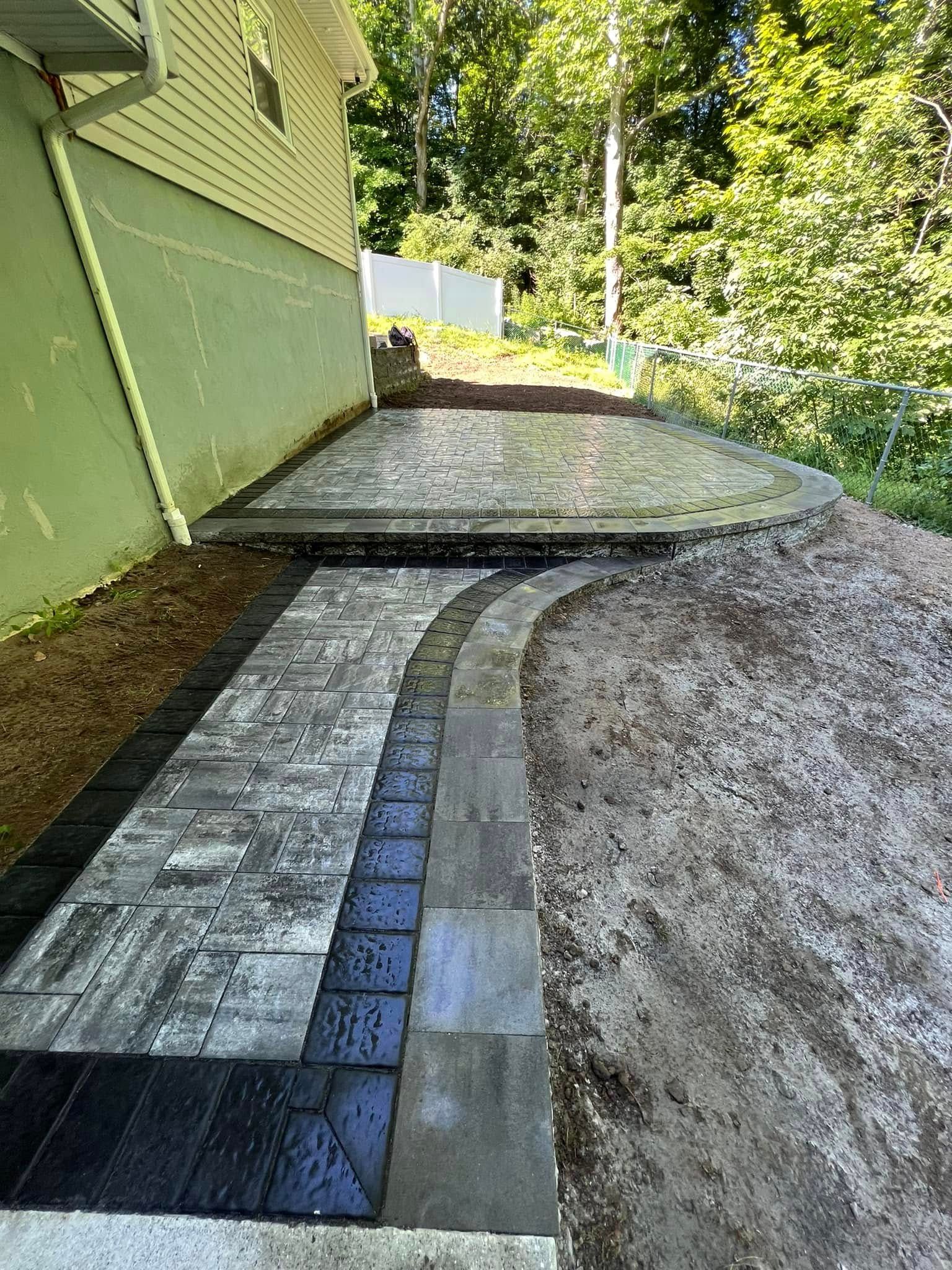 A paved walkway with dark border stones leads to a patio area adjacent to the light-colored exterior wall of a house.