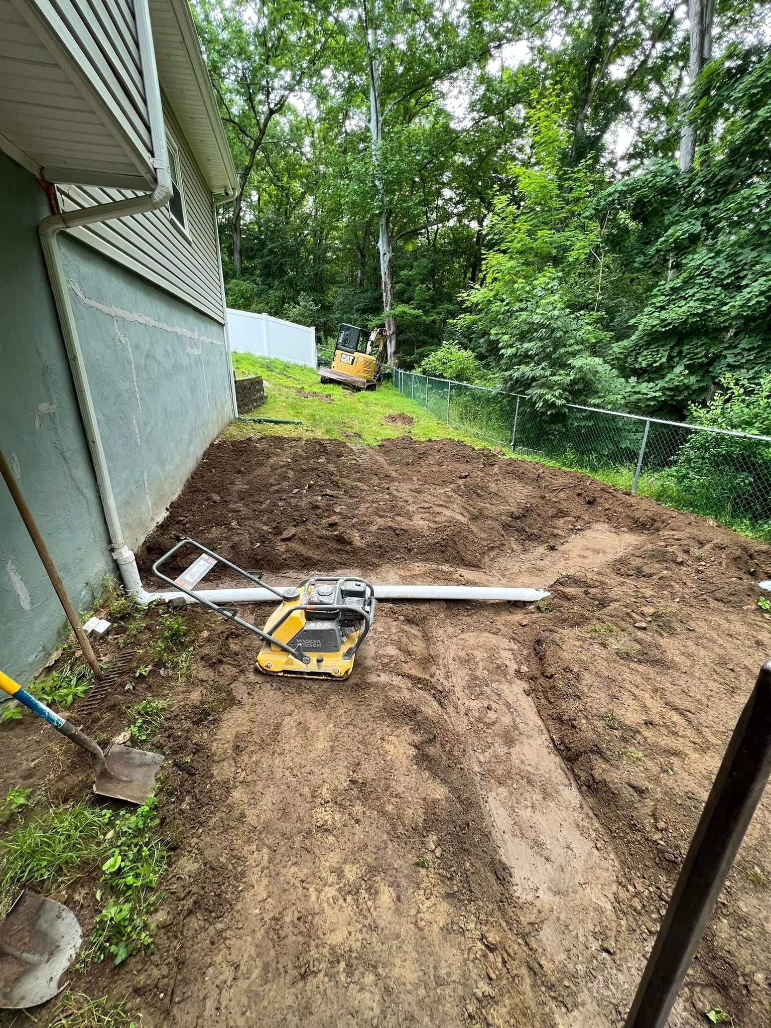 A residential yard undergoing construction, featuring a yellow plate compactor on fresh soil next to a house wall.