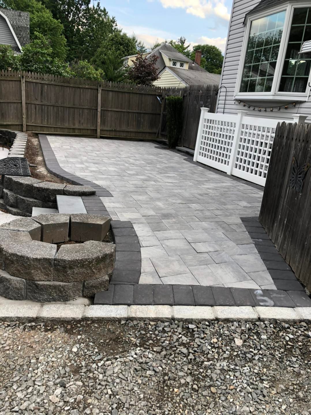 A light gray stone patio with dark gray border pavers, a stone fire pit, and a white lattice fence next to a house.