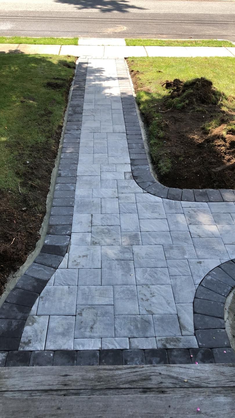 A grey paver walkway with a dark charcoal border extending from a wooden step toward a sidewalk and lawn.