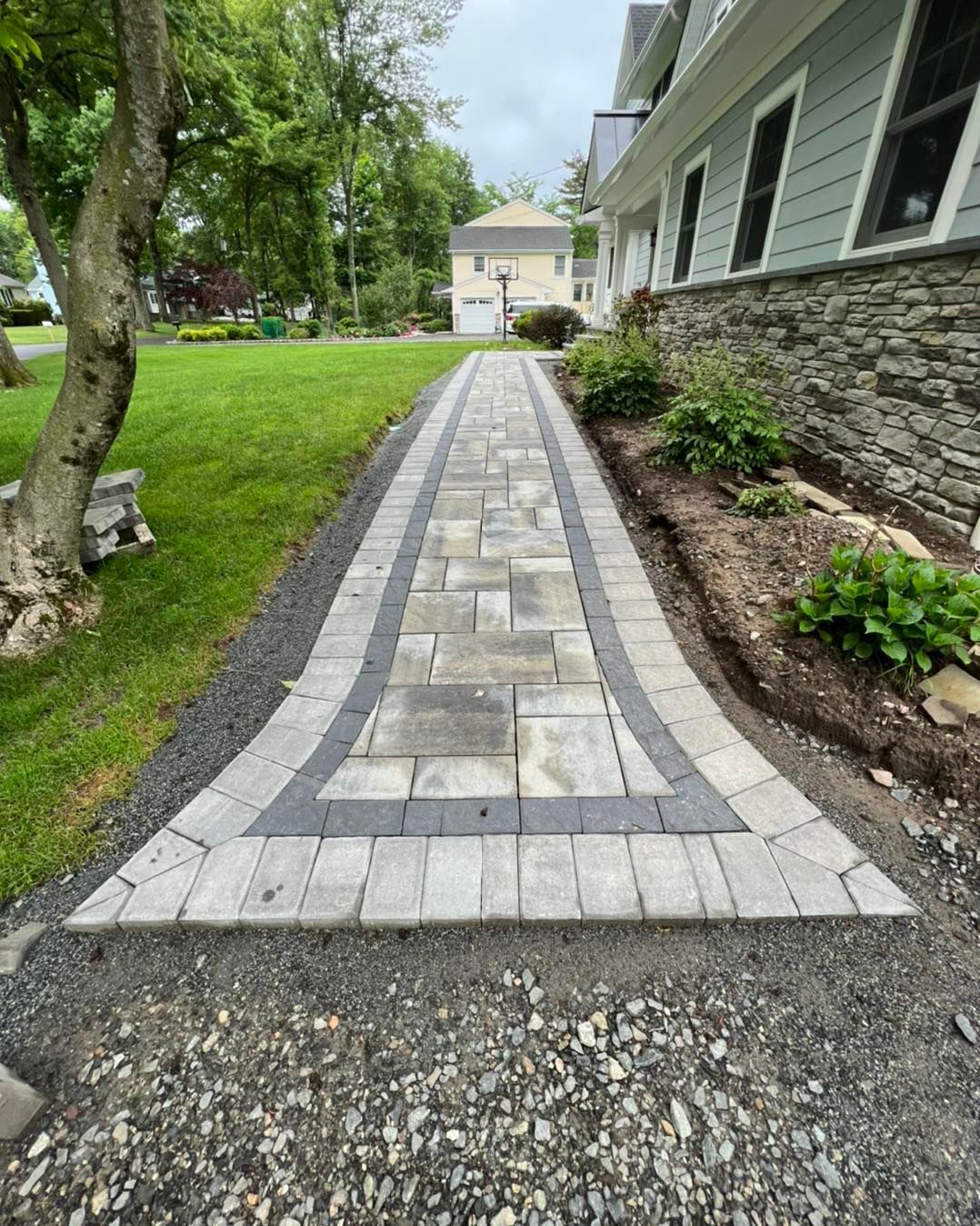 A paved stone walkway leads toward a house, bordered by dark stones, lush green lawn, and garden beds along the exterior.
