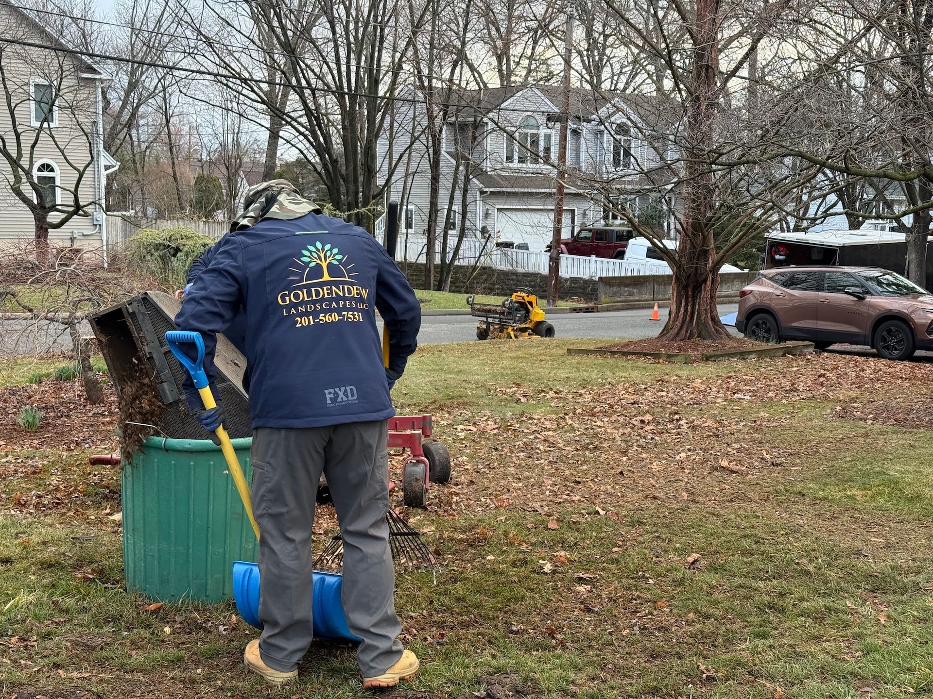 A person wearing a blue long-sleeved shirt is raking fallen leaves into a green bin in a suburban yard.