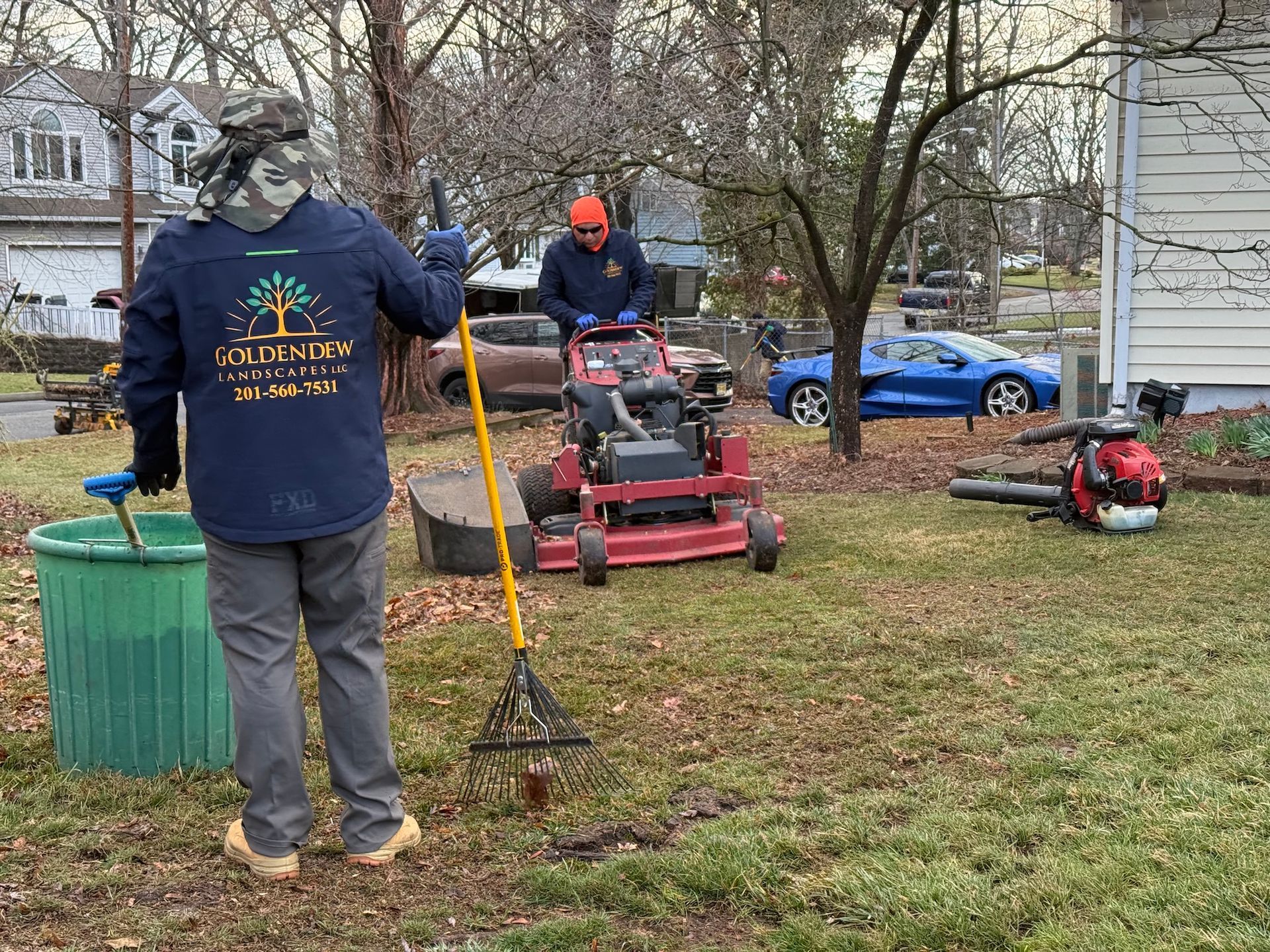 Two workers perform lawn care in a yard with a red riding mower, rakes, and a blue car parked in the background.